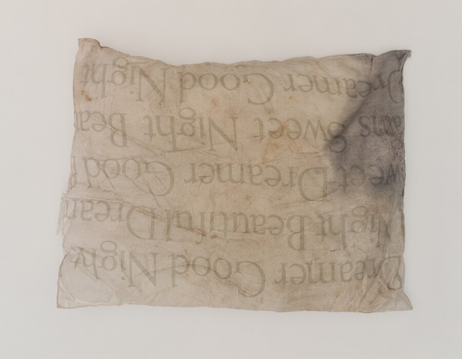 This is a photograph of a pillow with printed text. The image shows a slightly worn pillow with faint, greenish text phrases such as "Sweet Dreams" and "Good Night." The pillow has a crumpled appearance with some discoloration, particularly in the top right corner. The texture of the fabric is patchy and aged, and the background is plain and neutral, highlighting the pillow.