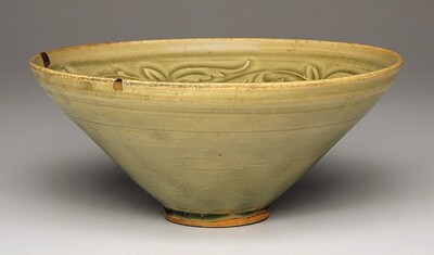 This is a photograph of a ceramic bowl. The bowl has a soft olive-green glaze with broad curving lines forming an abstract floral or vine-like pattern incised into the interior. The shape is wide at the top, tapering down to a small raised foot. The rim has small aged chips revealing a darker clay base. Subtle crackling is visible throughout the glazed surface, giving it an antiquated appearance. The lighting highlights the texture and color variations, accentuating the bowl's simple form.