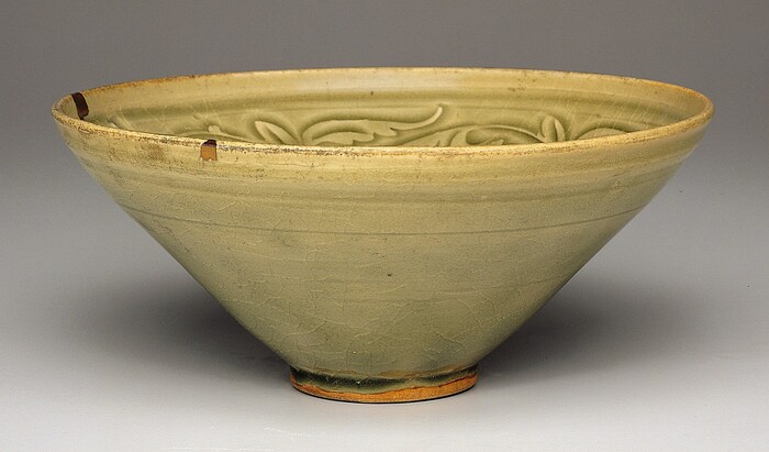 This is a photograph of a ceramic bowl. The bowl has a soft olive-green glaze with broad curving lines forming an abstract floral or vine-like pattern incised into the interior. The shape is wide at the top, tapering down to a small raised foot. The rim has small aged chips revealing a darker clay base. Subtle crackling is visible throughout the glazed surface, giving it an antiquated appearance. The lighting highlights the texture and color variations, accentuating the bowl's simple form.