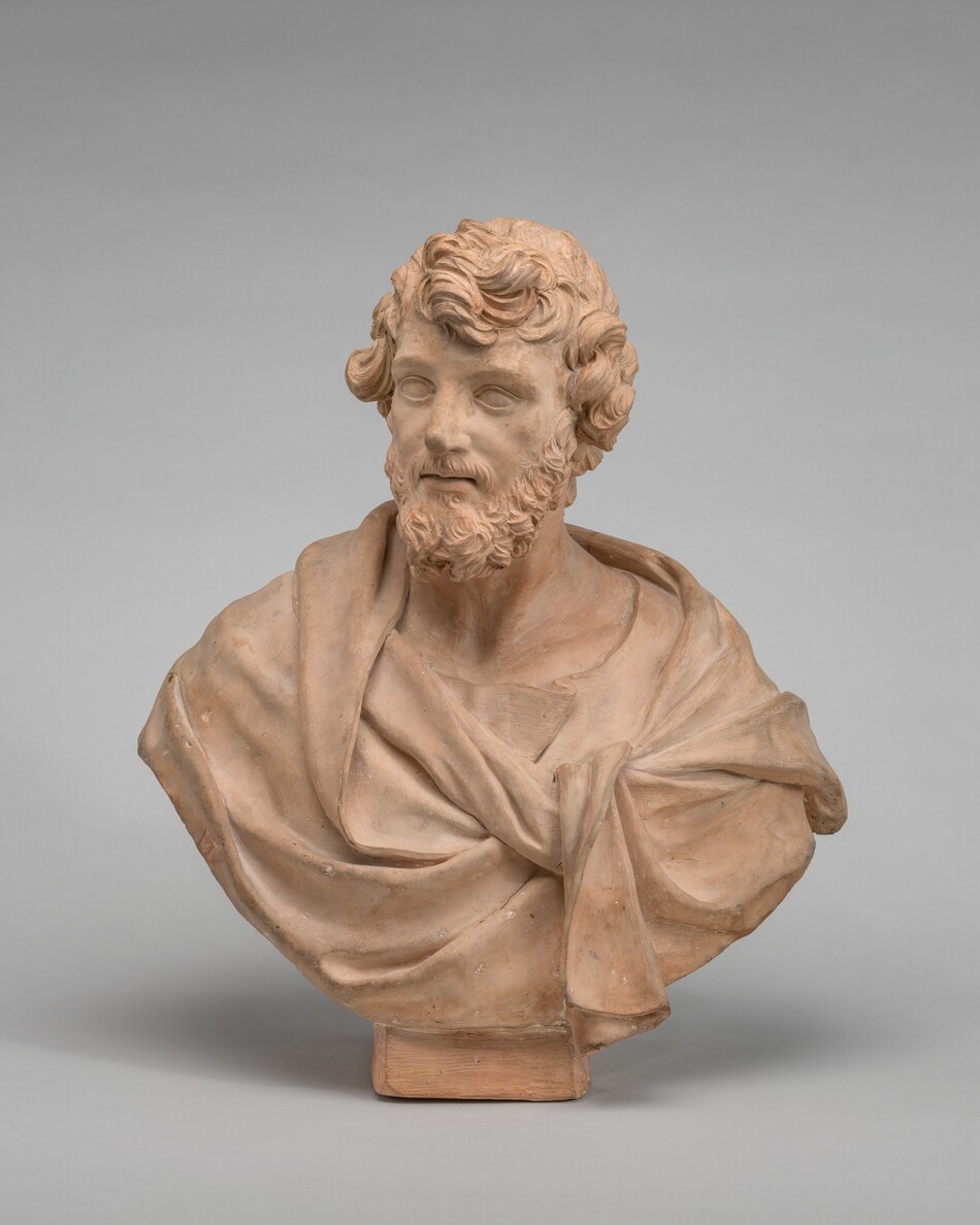The sculpture is a bust of a bearded man with curly hair, draped in a garment knotted at the front. It is made of terracotta, with a warm, reddish-brown color. The figure is detailed and lifelike, including features like the man's curls and facial hair. The eyes of the sculpture lack detailed pupils, giving it a classical and somewhat stylized appearance. The bust is presented on a simple rectangular base.