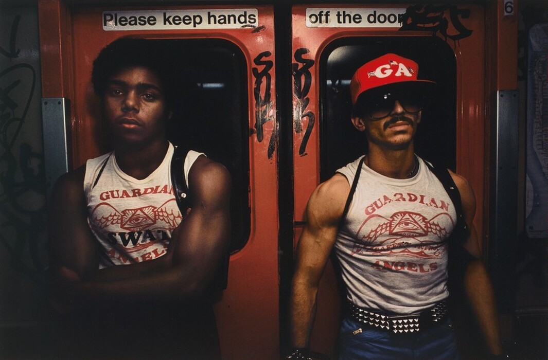 Two men are seen from the waist up, standing side by side near a subway door. The man on the left has short, curly hair and a shirt that reads "GUARDIAN ANGELS SWAT." The man on the right has a mustache, wears a red baseball cap with "GA" letters, and a shirt that also reads "GUARDIAN ANGELS." They are in a subway car with orange doors and graffiti.