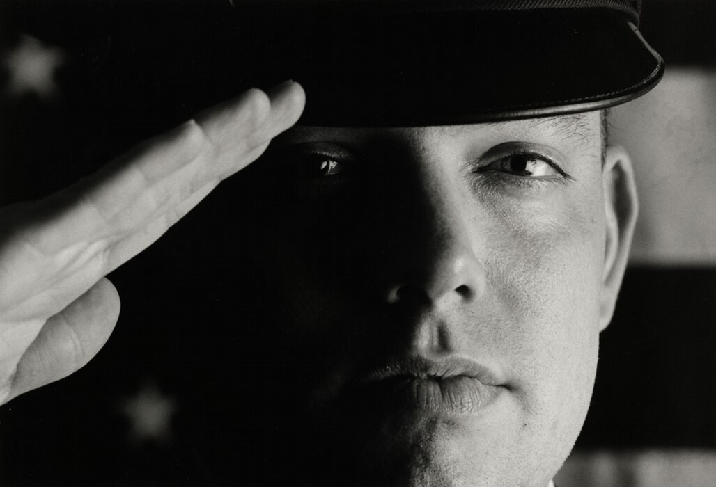 The image displays a close-up of a person's face and hand, with the focus on the face. The person is shown in a salute position, with their hand raised to their forehead. The facial features show a slight expression of concentration, an attentive and focused eye, and full lips. The person is wearing a dark-colored hat, which is part of their uniform. The background contains blurry shapes hinting at the presence of an American flag with stars and stripes. The lighting highlights the person's face against the background.