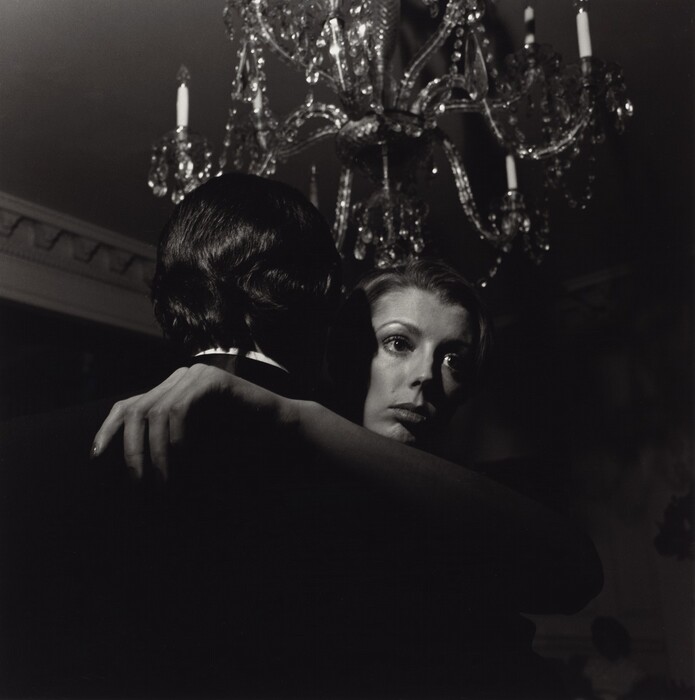 The image shows a man and a woman in an embrace, focusing on the woman's face and upper body. The man faces away from the camera, with his well-groomed, dark hair. The woman's left arm is draped over the man's shoulder, with her styled, light-colored hair swept back, and her facial features pronounced. She is looking towards something out of the frame. She is wearing a formal outfit, and the backdrop includes an ornate chandelier, decorative crystals, and architectural details.