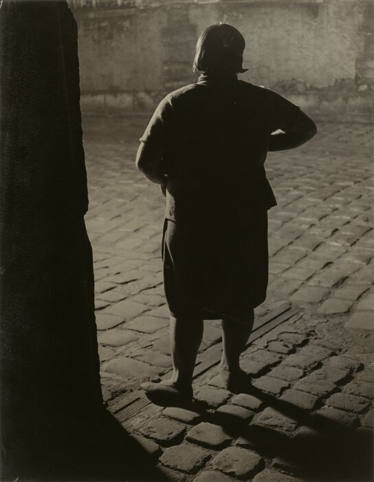 The image depicts a person standing with their back facing the camera. They are wearing a dark, structured top and a knee-length skirt. The individual's right hand is resting on their hip, while their left arm hangs by their side. They have short, dark, straight hair and their feet are cut off at the bottom of the frame. The background shows a cobblestone street or courtyard with shadows indicating a light source in front of them. In the background, there is a textured, possibly aged, wall with some geometric details at the top edge. The setting is outdoors and could be in the evening or nighttime.