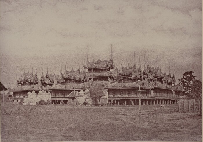 The image shows an architectural structure viewed from ground level. The building is a traditional palace with ornate, tiered roofs and intricate carvings. The horizon is low in the frame, emphasizing the roofline which stretches horizontally. The sky is overcast with soft clouds, casting subtle shadows. The image has a historical appearance, possibly with a sepia tone. It is dominated by shades of brown and grey, enhancing the vintage feel. The surroundings are simple, with posts and pathways leading to the building. Trees can be seen in the distance on either side, framing the architectural silhouette against the sky.