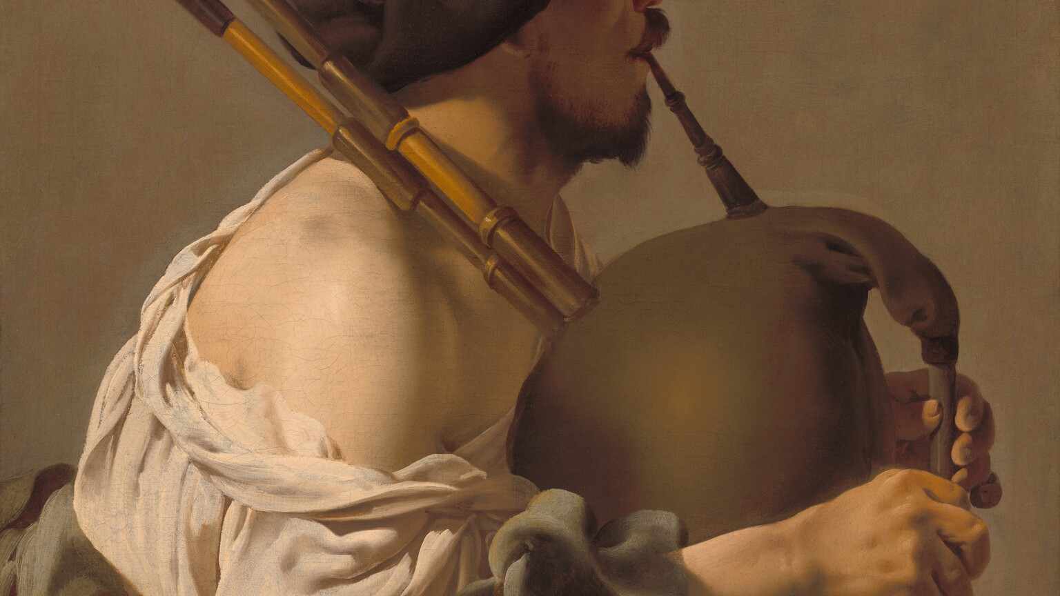 Shown from the lap up, a man with pale skin sits facing our right in profile, playing a bagpipe in this vertical painting. He is lit from our left so his face is in shadow, and he looks off to our right. He has dark eyebrows, a chestnut-brown mustache and short beard, and a rounded nose. He wears a floppy, chocolate-brown beret that covers his hair and the ear facing us. A cream-white, voluminous shirt falls off the shoulder close to us, and a fawn-brown robe gathers around his lap. He blows into a long mouthpiece as he squeezes the tawny-brown bag of the instrument between his forearms. Two long, wooden pipes rest over his shoulder, and he covers the finger-holes of the flute-like chanter at the front of the bag. The background behind him is pale peanut brown. The artist signed and dated the painting just to the right of the musician’s face, “HTBrugghen fecit 1624,” with the HTB intertwined into a monogram.