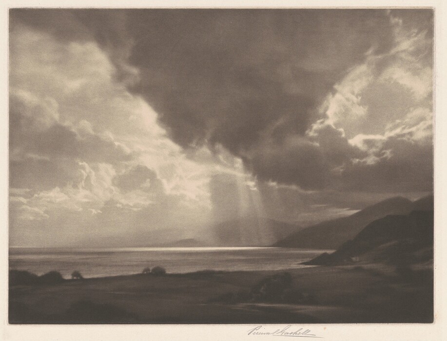 The image displays a vast body of water with land in the distance. The horizon is low, emphasizing the expansive sky filled with dense clouds and some sunlight. The water and landforms are partially lit, with hills or mountains fading into the distance. The foreground shows dark ground, potentially grassy or barren. The color scheme is sepia, creating a soft and timeless atmosphere. The tones are smoothly blended with a focus on light and shadow.