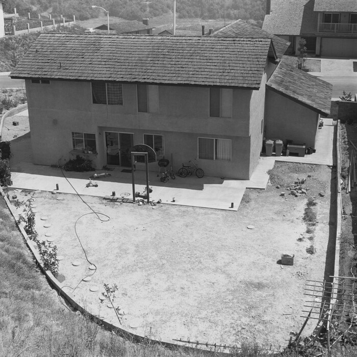 This is a photograph of a two-story house with a backyard area. The image shows a house with a shingled roof and several windows, some with closed blinds and others with curtains. Below the windows is a paved patio area, partially occupied by two bicycles, a tricycle, a barbecue grill, a small round table, and a few scattered outdoor toys. The patio leads to a largely barren dirt backyard enclosed by a low fence. There are a few plants and stepping stones along the edge of the yard, and a garden hose is loosely coiled on the ground. In the foreground, the yard slopes gently, and the dry earth is marked with patches of grass and sparse vegetation. The setting has an exposed, unfinished feel.