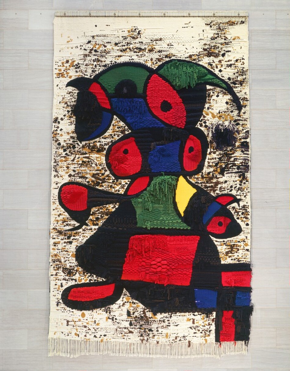 The abstracted form of a woman is made with blocks of cherry red, pine green, cobalt blue, black, and one canary yellow-shape on this vertical, woven tapestry. The ivory-white background of the tapestry is dotted and streaked with light and dark brown and black, and it’s fringed along the bottom. The colorful shapes roughly make up the form of a person with a headdress, arms, one foot, and perhaps a skirt. A grid of red, blue, and black blocks are also in the lower right corner. The weaving is not uniform, and ends hang down in many areas. The texture varies from fine, uniform stitches to scale-like sections. The artists’ names are woven into the tapestry near the middle of the right edge, “Miró” and “Royo.”