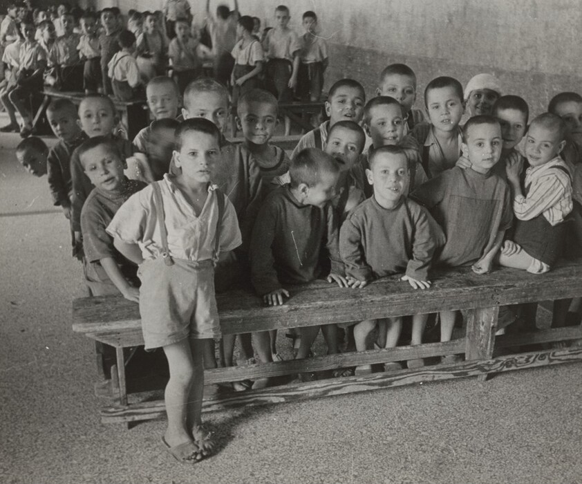 This is a photograph of a group gathered indoors. The image shows a large assembly of individuals arranged on wooden benches and standing against a plain, textured wall. The individuals have closely cropped hair and are dressed in simple attire, consisting of shorts, suspenders, and shirts. The setting appears to be a communal space, as indicated by the rows of benches and the tiled or concrete flooring. The lighting is natural and soft, casting gentle shadows throughout the room.