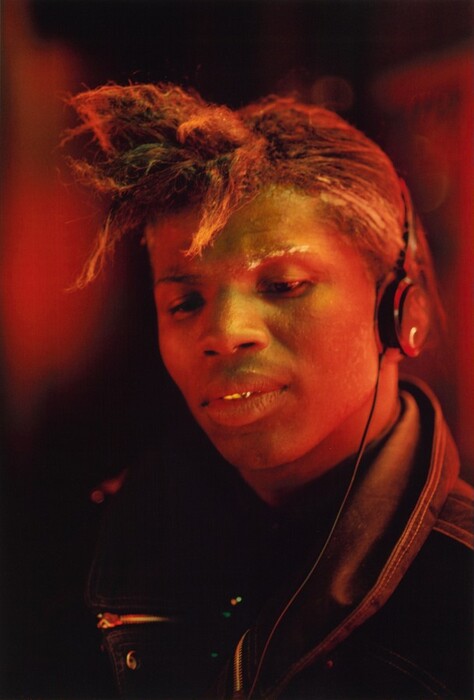 The image displays a close-up of a person, focusing on their head and shoulders. The person is gazing towards the camera with a unique hairstyle featuring thick, textured hair styled upwards. They have high cheekbones, full lips, natural eyebrows, and a relaxed facial expression. The person is wearing headphones with a visible wire and a black jacket with buttons. The background is blurred with warm red tones, creating a vibrant atmosphere around the subject.