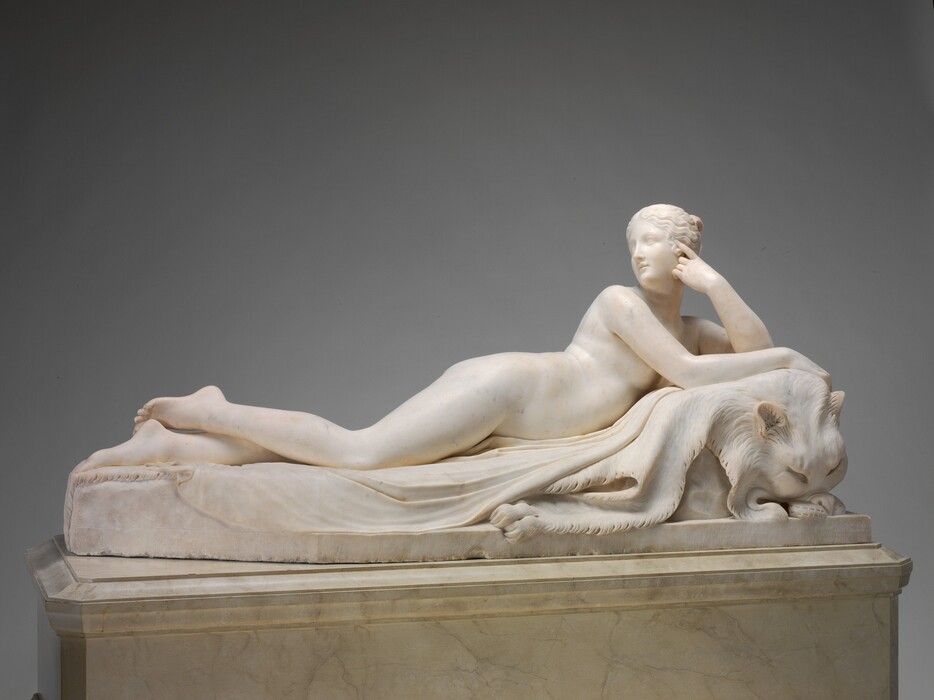 Carved from white marble, a nude woman lies belly down but tipped onto one hip and propped up on her elbows in this freestanding sculpture. In this photograph, the woman’s feet are to our left and her head to our right. Her upper body is further bolstered by what appears to be an animal skin – the flaccid hide of a lion, perhaps. Her right arm, closer to us, lies along the animal’s head, and her other elbow is bent so that pointer finger touches the side of her head. She turns sharply to look back over her right shoulder. She has delicate features, and her wavy hair is pulled back and up. There are two wrinkles where her waist cinches together as she turns to look to our left. One foot is daintily crossed over the other, and she lies on a cloth. The animal skin and cloth drape over a slab-like base. The polished, chest-shaped marble plinth is parchment white with gray veining, and the sculpture is photographed against a gray background.