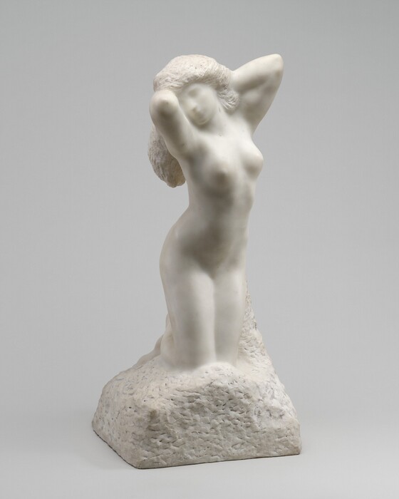 A woman kneeling with both arms raised by her head is carved from white marble in this freestanding sculpture. In this photograph, her body is angled slightly to our right and her face turns toward us. Her face and body are smooth and carved so her features appear soft and indistinct. Both elbows are raised by her long, textured hair as if caught mid-stretch. Chisel marks are also visible on the base, which appears to be a rounded boulder on which she kneels.