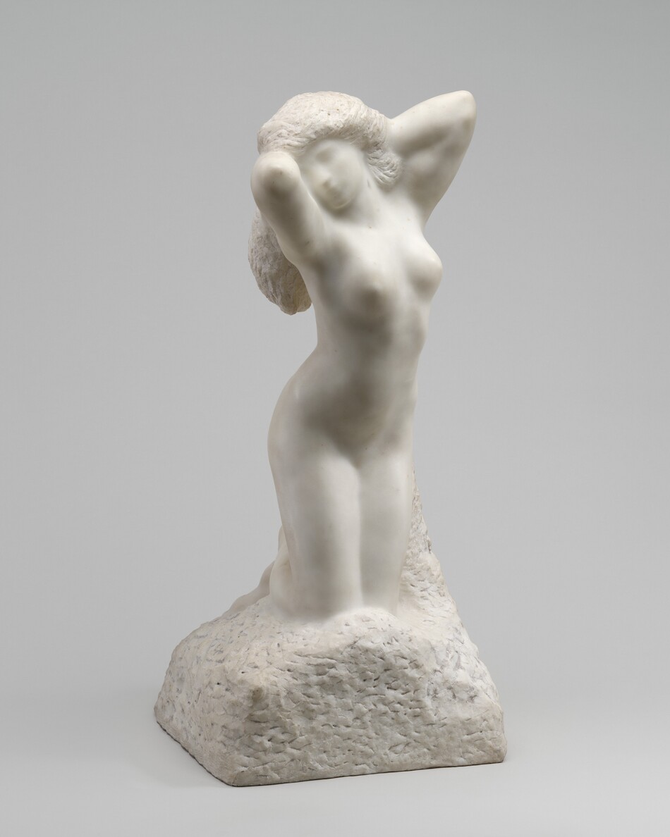 A woman kneeling with both arms raised by her head is carved from white marble in this freestanding sculpture. In this photograph, her body is angled slightly to our right and her face turns toward us. Her face and body are smooth and carved so her features appear soft and indistinct. Both elbows are raised by her long, textured hair as if caught mid-stretch. Chisel marks are also visible on the base, which appears to be a rounded boulder on which she kneels.