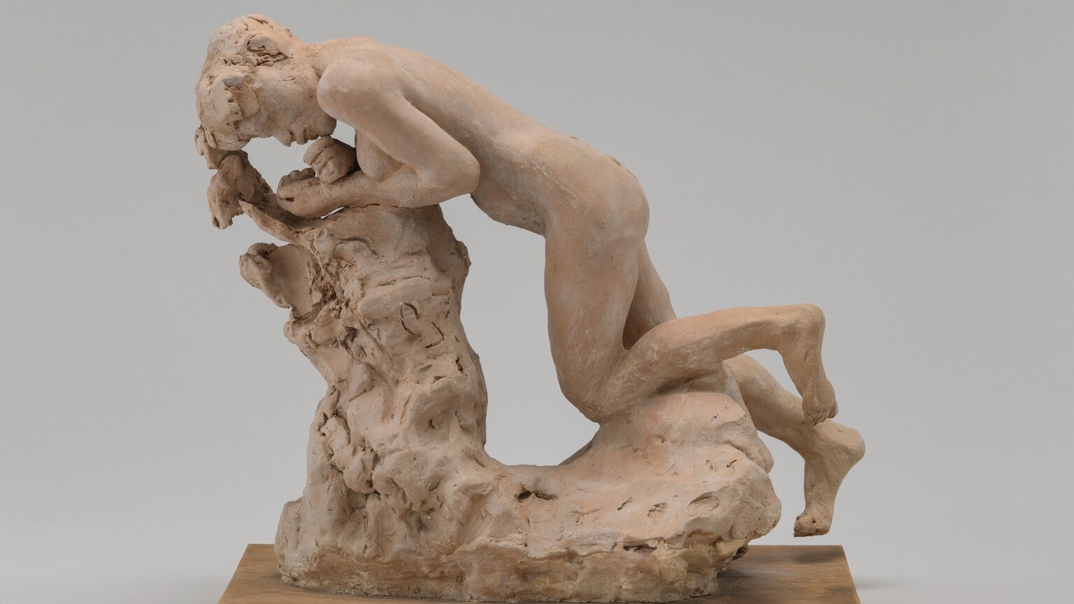 Molded in tan-colored terracotta, a person kneels and leans forward against an L-shaped stump-like form in this freestanding sculpture. The person’s head is to our left in this view, and the breasts are visible. She leans forward onto her forearms with her wrists crossed, so her hands are near her chin and closed mouth. She has short hair and full lips. The area around her eyes is loosely sculpted. One knee is bent and the other is mostly straight as if the foot were reaching toward the ground. The stump is especially loosely formed, and the sculpture sits on a wooden plank. The piece is photographed against a gray background.