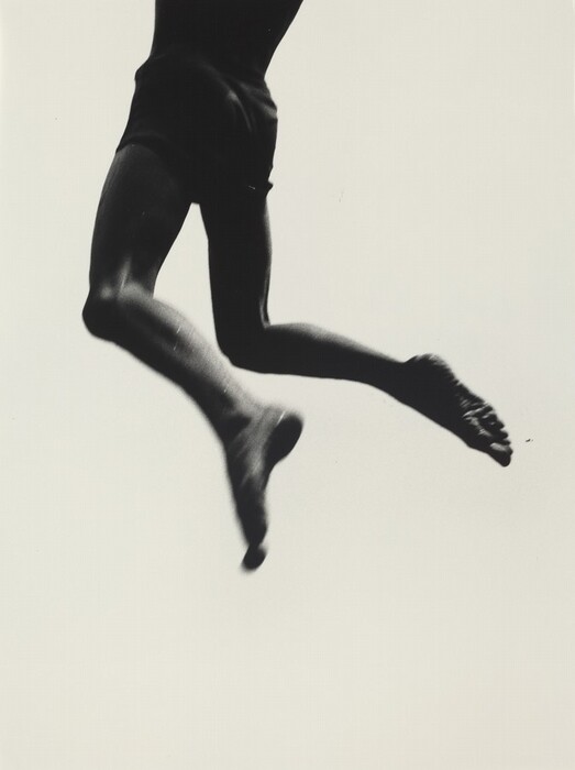 This is a black and white photograph of a person jumping, capturing their legs and torso. The image emphasizes the contrast between the figure's silhouette and the light background. The focus is on the dynamic movement and graceful posture of the subject's leap, with one knee bent and the other leg extended, and their feet slightly pointed. The photograph conveys a sense of motion and fluidity, capturing a moment of athletic elegance. The high contrast highlights the musculature and form against the stark background, drawing attention to the composition's simplicity and the expression of movement.