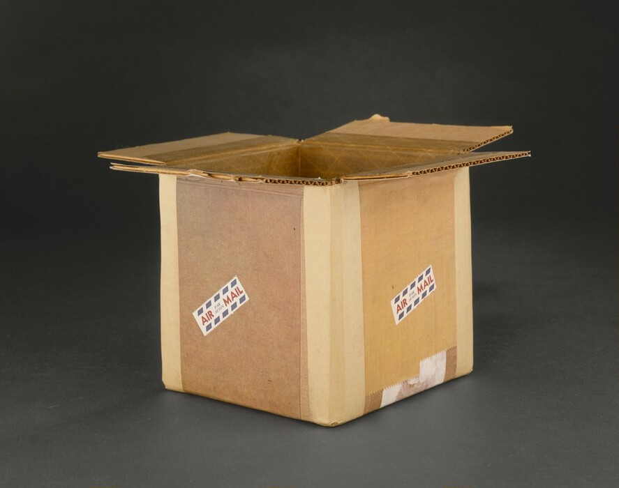 The sculpture depicts a cardboard box with its flaps open. It is brown and appears to be made of cardboard material. There are two small labels on its side with text and design reminiscent of "AIR MAIL" stickers. The box is placed slightly tilted.