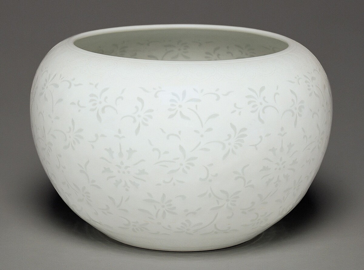 The sculpture is a large, rounded bowl with a delicate floral pattern covering its smooth surface. It is predominantly white, with the floral motifs painted in a light gray or off-white. The bowl has a wide, open top, and the same painted pattern appears to continue inside the bowl.