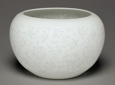The sculpture is a large, rounded bowl with a delicate floral pattern covering its smooth surface. It is predominantly white, with the floral motifs painted in a light gray or off-white. The bowl has a wide, open top, and the same painted pattern appears to continue inside the bowl.