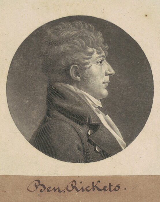 The image shows a portrait of a man in profile view. He has distinct facial features, including a prominent nose and defined lips. His hair is styled in thick, wavy curls. He is dressed in formal attire with a dark coat and a light-colored cravat tied around his neck. The background is plain and dark. The portrait is set within a circular frame, with the handwritten name "Ben. Rickets" below it.