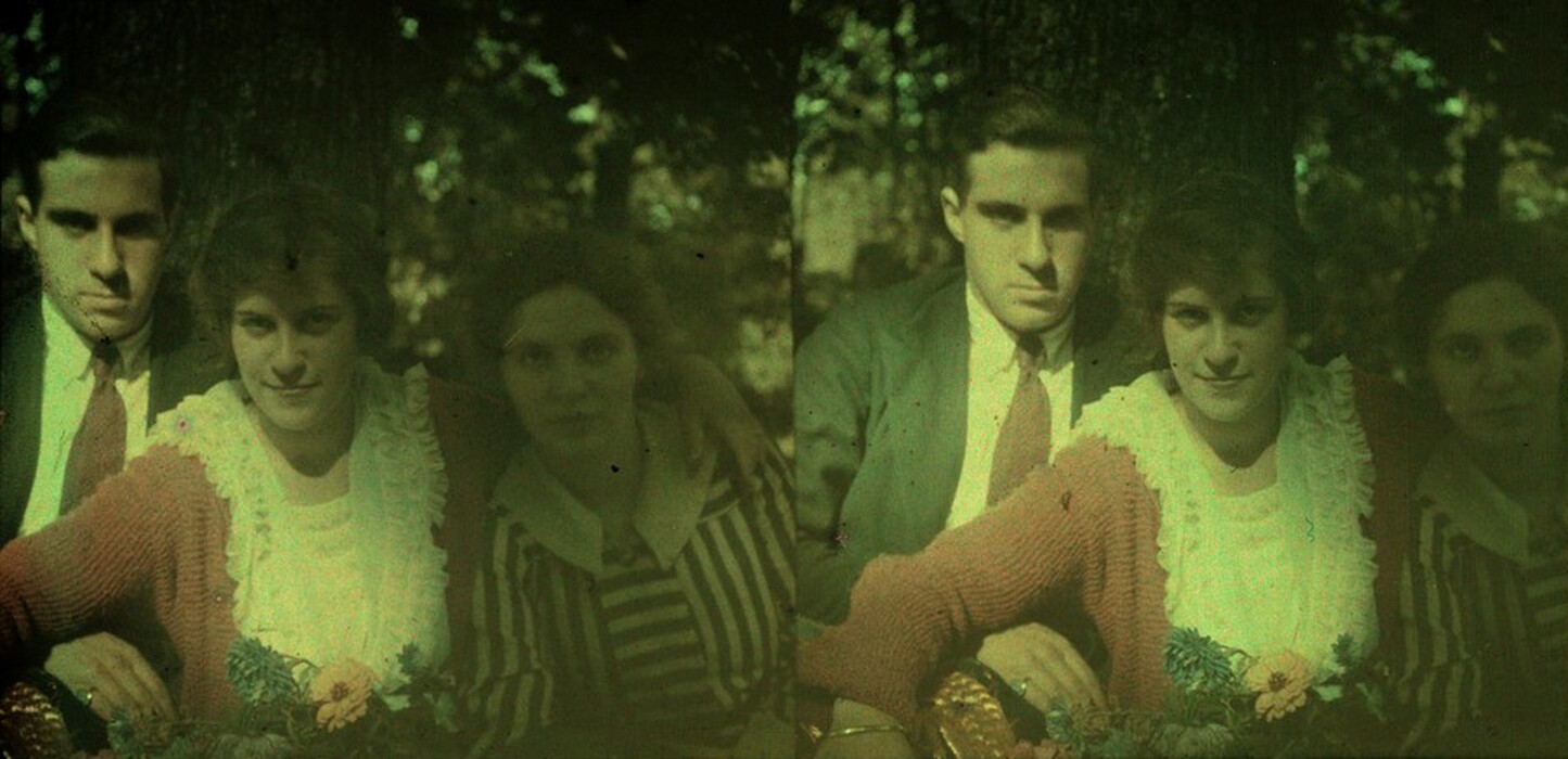 The image shows three individuals sitting closely together. The person on the left is a man with dark hair, wearing a dark suit jacket, a white shirt, and a red tie. In the center, a woman with short, wavy hair is wearing a red knitted cardigan over a white ruffled blouse. To the right, there is another woman with dark hair, wearing a vertically striped blouse with a white collar. The first woman appears to have her arm around the second woman, with a floral arrangement in the foreground. They are seated outdoors with a large tree trunk behind them, and the dappled light through the leaves creates a shaded, rustic ambiance in the background.