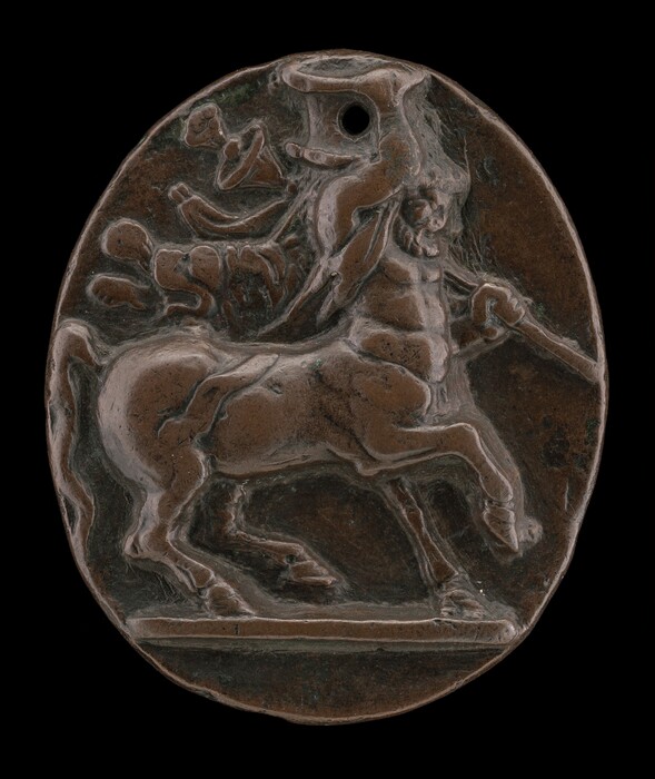 The sculpture shows a centaur rearing on its hind legs. It is made of dark brown material, possibly bronze, and features shallow relief work for detailing. The centaur holds a long stick or staff and exhibits animated facial features. The sculpture is set on an oval background with a hole at the top and decorative swirling patterns around the centaur.