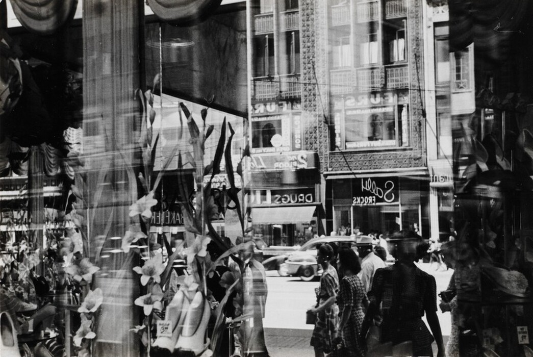This is an image of a city street reflected in a shop window. The scene includes decorative elements like flowers and merchandise in the foreground, with historical buildings and shop signage visible on the street behind. There are cars and blurred pedestrians in the background, showcasing a dynamic urban environment.