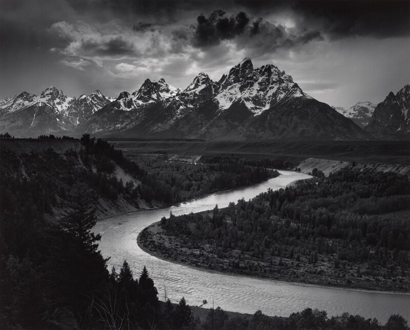 We look down into a darkened valley with a shimmering, curving river winding back to a screen of craggy, snow-topped mountains in the distance in this horizontal black and white photograph.