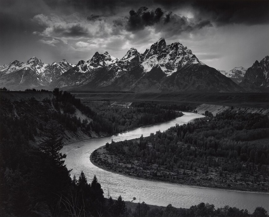 We look down into a darkened valley with a shimmering, curving river winding back to a screen of craggy, snow-topped mountains in the distance in this horizontal black and white photograph.
