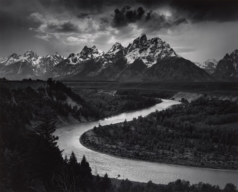 We look down into a darkened valley with a shimmering, curving river winding back to a screen of craggy, snow-topped mountains in the distance in this horizontal black and white photograph.