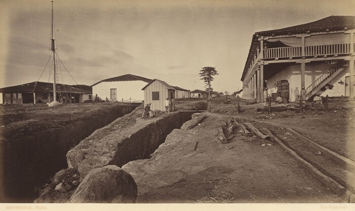 The image shows an aerial view of a landscape with a trench-like excavation extending into the distance. Buildings with dark, sloping roofs are aligned parallel to the trench on the left side. A tall mast with a ladder is next to the nearest building. On the right, a larger two-story structure with a covered porch supported by wooden posts is visible. Several people are present in the scene, standing near the buildings or sitting by the trench. The ground is barren, with scattered logs and rocks. The color palette is monochromatic with sepia tones.