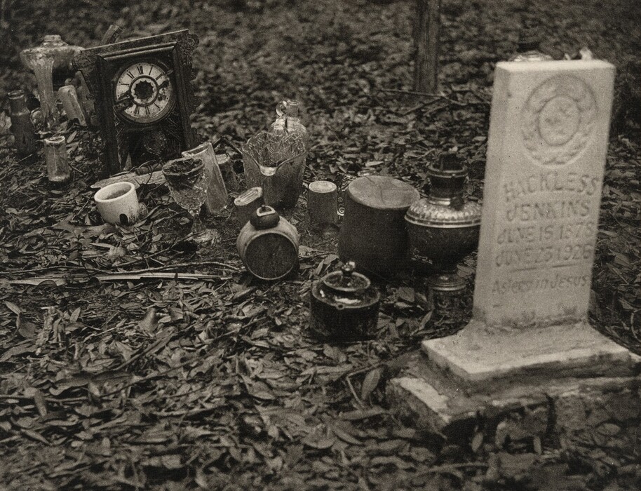 There is a gravestone on the right side of the image. On the left side, there are cups, bottles, an oil lamp, and a clock. The color palette is muted, with gray and beige tones, creating a sepia effect. In the foreground, there are glass and ceramic objects scattered on the ground with fallen leaves.