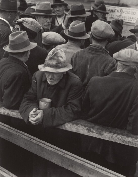 More than a dozen men crowded together fill this vertical black and white photograph. All wear snap-brim hats, fedoras, or caps, and all but two away from us. One man in the crowd looks to our right. The other is closest to us. He rests his forearms on a wooden railing, a metal cup tucked between his elbows. His hands are tightly clasped, and his worn, stained fedora is low over his lined face. His thin lips are set and he has a grizzled white beard. A partially legible sign in the back reads "SAN FRANCISCO" and "BY THE GOLDEN GA."