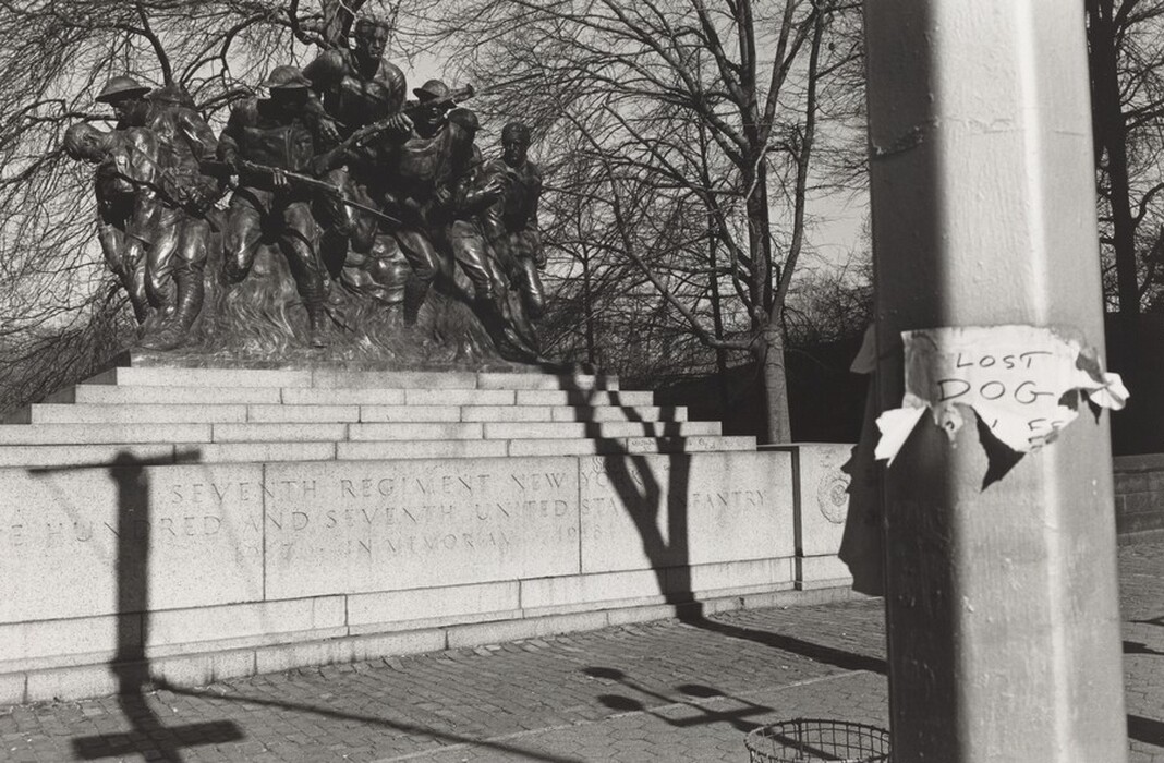 The sculpture depicts a group of soldiers engaged in battle or advancing together. The sculpture is made of dark material, likely bronze, and sits on a stone base. The soldiers are shown with detailed uniforms, appearing determined and in motion. The inscription on the base mentions the "Seventh Regiment New York" and "United States Infantry." The soldiers are seen from a slightly side perspective. Beside the sculpture is a pole with a weathered 'lost dog' sign attached.