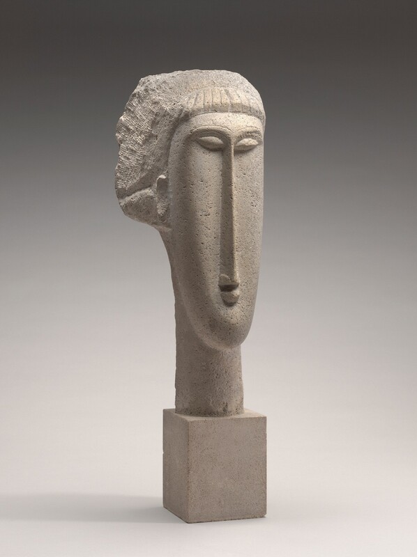 The elongated head and neck of a person with stylized features is carved from beige-colored limestone for this freestanding sculpture. Though mostly carved smooth, the surface of the porous limestone is pocked and textured. In this photograph, the face is angled to our right. Short bangs line the narrow forehead of the tall, oval face. The hair flaring around the crown and behind the tidy, oval ear we see is roughly carved. The marquis-shaped eyes are set high on the face. Eyebrows immediately over the eyes join to make a long, blade-like nose that nearly reaches the bottom of the oval face. The arrow-shape of the nose ends just above a half-moon-shaped lip over a round chin. The long neck continues to a block of limestone that acts as the base. The sculpture is photographed in front of a background that lightens from charcoal gray across the top to nearly white across the bottom.