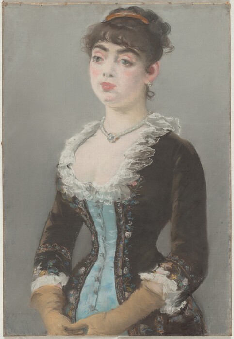 Shown from the hips up, a pale-skinned woman wearing a tightly corseted dress looks off to our left in this vertical pastel drawing. The portrait is loosely sketched with blended areas, giving it a soft look. The woman’s body is angled to our left, and her hands touch in front of her lower belly. She looks off with large, wide set, slightly bulging green eyes under dark brows. She has a round face, full pink lips, and the suggestion of a dimple in her chin. Her dark brown hair is pulled up under a gold band, and bangs curl across her forehead. A short, delicate ringlet curls up the nape of her neck. She wears a light-colored, round earring in the ear we see and a string of pearls, which may hold a round jewel that rests in the hollow of her throat. Her dress is powder blue down the front and coffee bean brown to either side and on the sleeves. The edges of the brown sections are lined with flowers drawn loosely in shades of pastel pink, blue, yellow, and green. The low, curving neckline is edged with white ruffles, and the waist is cinched to an almost startling degree. The sleeves come down just past her elbow, and the cuffs are also lined with white ruffles. Long, tan-colored gloves stretch back to the cuffs. The background is fog gray. The artist signed and dated the pastel faintly in the lower left corner, “Manet 1882.”