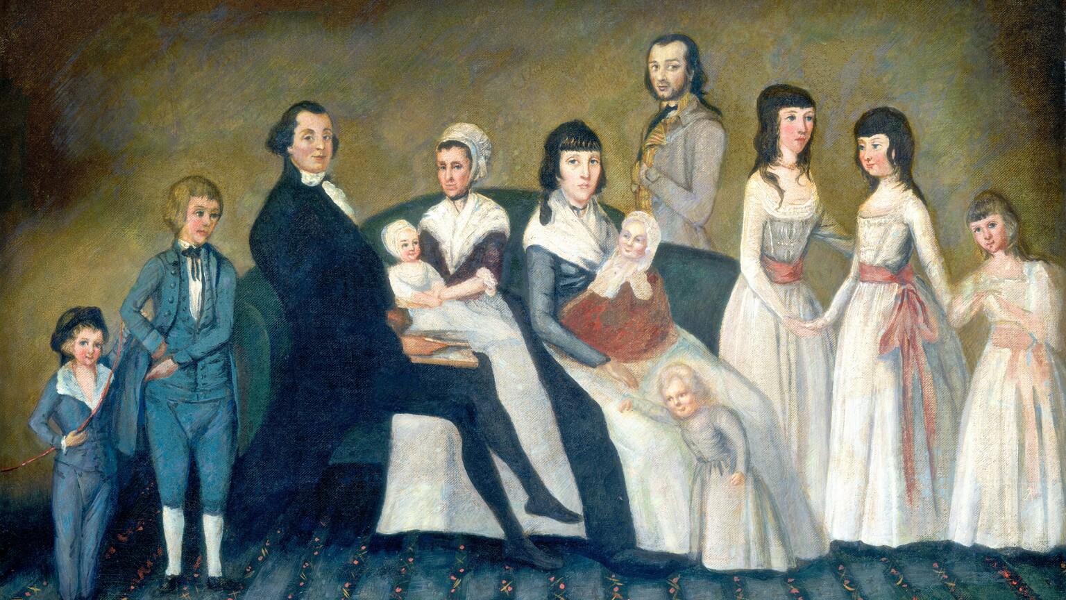 Twelve men, women, children, and babies are arranged on or around a couch in this horizontal portrait painting. The features and clothing are simply painted, so some of the faces are a bit generic. They all have pale skin with rosy cheeks, and all but one of them have blue eyes. The slate-blue couch is centered against a wall mottled with copper brown, golden yellow, and sage green. Three adults sit on the couch. To our left, a man wearing a black suit and stockings leans against the arm of the couch so his body faces our right in profile, his legs crossed. He turns to look at us with a slight smile on his lips, his eyebrows raised. His dark hair is smoothed back along the top of his head and is bushy over each ear. A white neckerchief wraps across his high collar and ruffles cascade down the front. He holds a book in his lap. Two women sit next to him, to our right. Both have dark hair and wear broad neckerchiefs over tightly fitting bodices, and white aprons over long, full skirts. The woman to our left turns her face slightly to our left but looks at us from the corners of her eyes. She wears a white bonnet, and her bodice is plum purple. The other woman’s head is uncovered and her forehead lined with bangs. Both of these women hold babies wearing white caps, and the babies smile. Just behind the couch, to our right of the second woman, a young man with a faint, dark beard, black hair, and brown eyes stands with his body facing our left in profile. He turns to look at us as he tucks one hand between the buttons of his nickel-gray, high-collared jacket. Two boys wearing blue suits stand to our left in front of the couch, near the seated man. The smaller boy, to our left, holds an archery bow, wears a dark cap, and looks at us, smiling. The older boy standing next to him has blond hair and points down at the younger child with one hand, the other on his hip. Two young women and a girl stand to our right, also in front of the couch. They all wear white dresses with long sleeves and skirts, their narrow waists tied with rose-pink sashes. Two older girls have black hair and hold hands. The youngest girl, near the right edge of the painting, has blond hair and also looks at us. The carpet beneath the group has vertical stripes leading back to the wall. Narrow bands of navy blue are dotted with pink to suggest a floral pattern, between wider, light blue stripes.