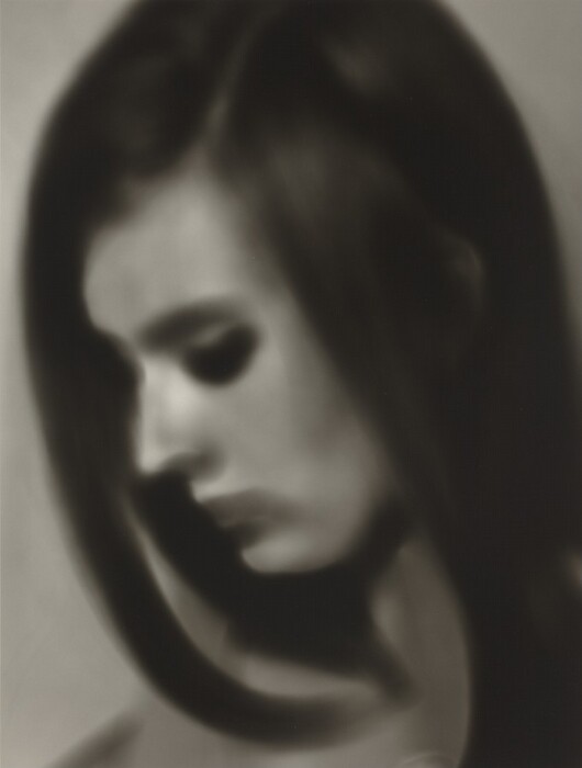 The image shows a person from the shoulders up. The person is in profile, facing slightly downwards. The facial features are blurry, with high cheekbones and a well-defined nose. The hair is dark, smooth, and straight, reaching past the jawline. The clothing is a neutral shade. The background is plain. No jewelry or objects are visible.