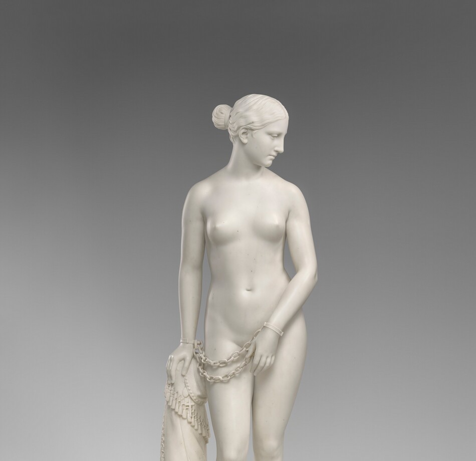 Carved from creamy white marble, a nude woman stands next to a hip-high support, perhaps a low post. In this photograph, her body faces us, and she looks down to our right in profile. Her wavy hair is tucked behind her ear and drawn back in a bun at the nape of her neck. Her weight rests on her left leg, on our right, and her other knee is bent. Her left arm is angled in front of her body so her hand covers her groin. Her other hand, on our left, rests on the post. Chains hang from shackles encircling her wrists. The post is covered with a cloth that gathers around the top and spirals to the ground beneath her feet, the edge trimmed with tassels. A cross and medallion peek out from under the cloth near her hand. She stands on a circular base.