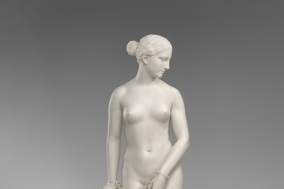 Carved from creamy white marble, a nude woman stands next to a hip-high support, perhaps a low post. In this photograph, her body faces us, and she looks down to our right in profile. Her wavy hair is tucked behind her ear and drawn back in a bun at the nape of her neck. Her weight rests on her left leg, on our right, and her other knee is bent. Her left arm is angled in front of her body so her hand covers her groin. Her other hand, on our left, rests on the post. Chains hang from shackles encircling her wrists. The post is covered with a cloth that gathers around the top and spirals to the ground beneath her feet, the edge trimmed with tassels. A cross and medallion peek out from under the cloth near her hand. She stands on a circular base.