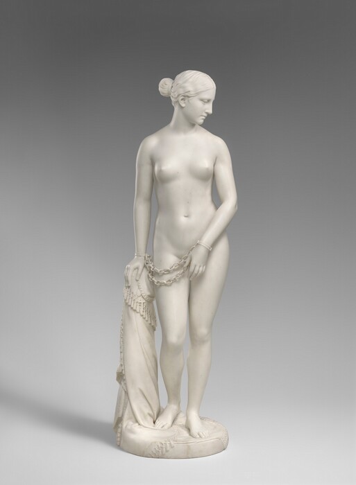 Carved from creamy white marble, a nude woman stands next to a hip-high support, perhaps a low post. In this photograph, her body faces us, and she looks down to our right in profile. Her wavy hair is tucked behind her ear and drawn back in a bun at the nape of her neck. Her weight rests on her left leg, on our right, and her other knee is bent. Her left arm is angled in front of her body so her hand covers her groin. Her other hand, on our left, rests on the post. Chains hang from shackles encircling her wrists. The post is covered with a cloth that gathers around the top and spirals to the ground beneath her feet, the edge trimmed with tassels. A cross and medallion peek out from under the cloth near her hand. She stands on a circular base.