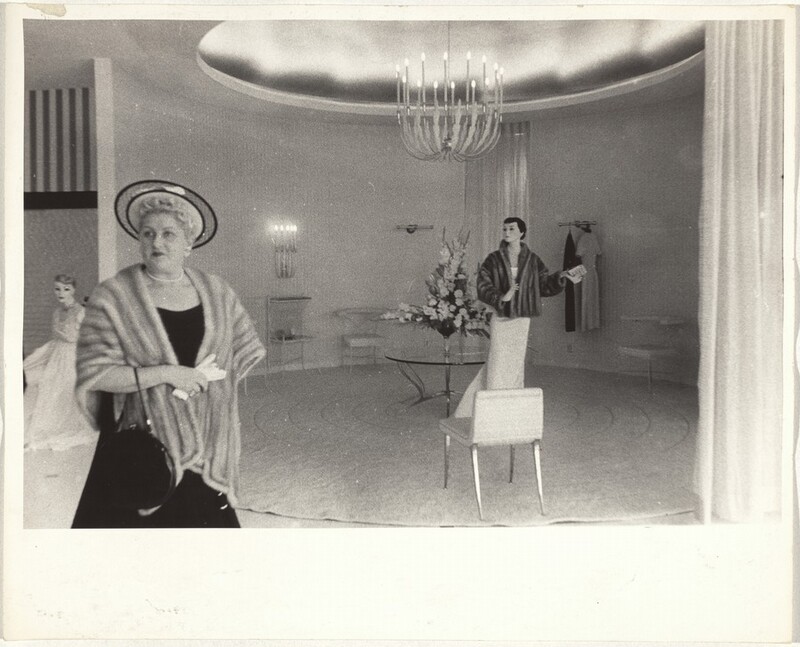 This is a photograph of a room with fashionable decorations. The image shows a well-furnished interior featuring a mannequin styled with elegant attire, including a fur coat. The room is spacious and circular with a patterned carpet and contains round-backed chairs and a central glass table with a floral centerpiece. A large, ornate chandelier hangs from the ceiling. The walls are adorned with subtle, decorative elements, and a pair of clothes hang on a wall-mounted rack. Additionally, a mannequin stands in the forefront, dressed in period attire.