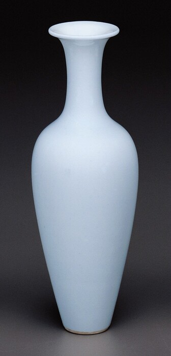 The sculpture is a blue vase with a tall, slender neck that widens into a bulbous body before tapering towards the bottom. The vase is made from smooth ceramic material and stands upright with a symmetrical shape, reflecting light.
