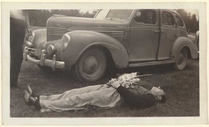 Untitled (Man lying on grass holding flowers in front of car)