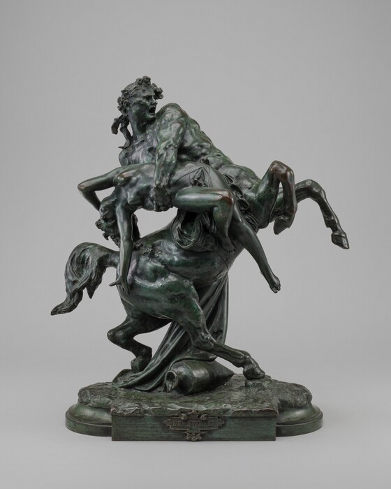 A centaur, a creature with a man’s torso and horse’s body, wraps both arms around a nearly nude woman who falls backward in this bronze, freestanding sculpture. In this photograph, the centaur faces our right with its front feet rearing up. His muscular torso leans to our left, and his mouth is wide open and brows deeply furrowed. A wreath of flowers is tied with a bow around his head, and he looks to the right. In his arms, the woman’s legs and arms fall back as she arcs away from him. One knee and one elbow are bent, and drapery covers only her groin. Her hair cascades onto the centaur’s rump, and her eyes and mouth are wide open. The centaur’s tail is lifted, and one back foot is planted forward, under the bulk of his body. The other leg is bent so the top of that hoof rests on a swath of fabric that falls down the far side of the pair. Water pours from an overturned urn onto a textured ground between the centaur’s feet. The cast base has molding and a plaque on the front reading, “L’ENLEVEMENT.” The bronze is shiny brown in some areas with a pine-green patina in other areas.