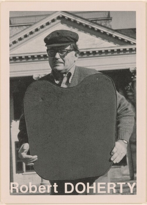 This is a photograph of a standing figure in front of a neoclassical building. The image is in black and white, featuring a person holding a large, dark-colored shield-like object in front of their chest. The figure is wearing a flat cap, glasses, a collared shirt, and a sweater. The background includes a neoclassical building with a pediment featuring dentil molding. At the bottom, the text "Robert DOHERTY" is displayed in white against a black band.