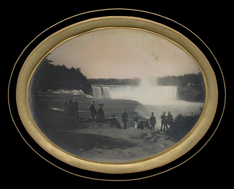 The image is a black-and-white photograph with a circular frame showing a group of people standing on a ledge by a large waterfall. The perspective is from behind the individuals, as they face the waterfall. Mist rises from the water, obscuring the horizon. The people are dressed in period attire and are silhouetted against the water. Vegetation is on the right, and trees line the distant riverbank.