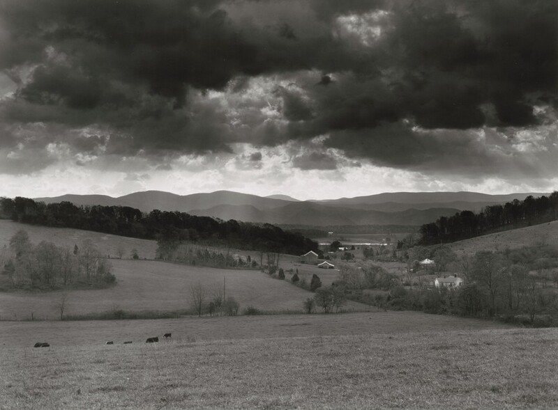 Hills roll gently back to a mountain range under a low sky dense with dark clouds in this horizontal black and white photograph. The grassy field closest to us leads back to a few dark cows grazing in a dip in the near distance. Lines and groves of lacy, spindly trees are farther back, surrounding a few structures and houses. Lines of darker trees creating a shallow V across the center of the composition are separated by a gap not far from the buildings. Strips of mountains are layered like torn, cut paper along the horizon, which comes halfway up this composition. Dark clouds roiling in the sky above are alleviated by a band of brighter sky along the mountaintops.