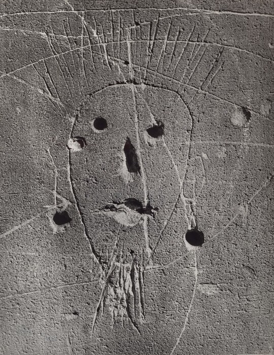 The image shows a carved representation of a human face on a textured surface. The face is depicted using basic lines and shapes, with two round indentations for eyes, an elongated indentation for the nose, and a line or space for the mouth. Lines suggest hair on top of the head. The background is a stone-like surface with scratches and imperfections. No other objects are depicted.