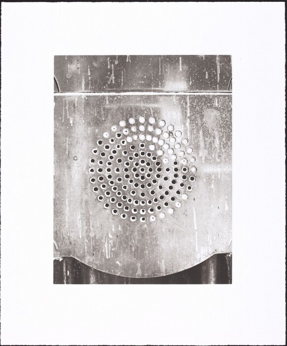 This is a photograph of a metallic surface with circular perforations arranged in a spiral pattern. The image captures a section of a metallic object or structure, with the central focus on a series of holes forming a spiral. The surface of the metal is weathered, showing signs of use or exposure to the elements. The overall tone is monochromatic, highlighting the texture and pattern of the metal surface against a clean, unobtrusive background.
